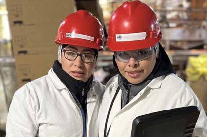 Two people wearing red hard hats and white lab coats stand in a warehouse, one holding a tablet.