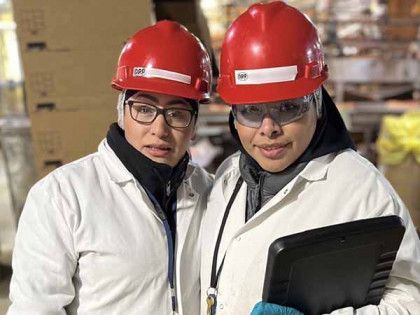 Two people in white coats and red hard hats, one holding a tablet, pose in an industrial warehouse setting.