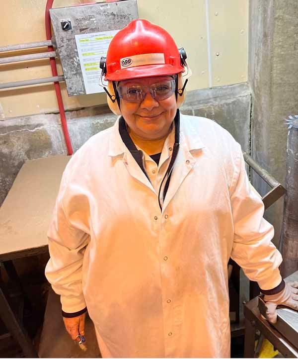 Person in a white lab coat, red hard hat, and safety glasses stands in a workspace, smiling at the camera.