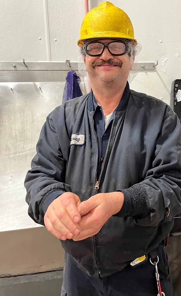 A person wearing a yellow hard hat, safety glasses, and a dark work jacket, smiling while rubbing their hands together.