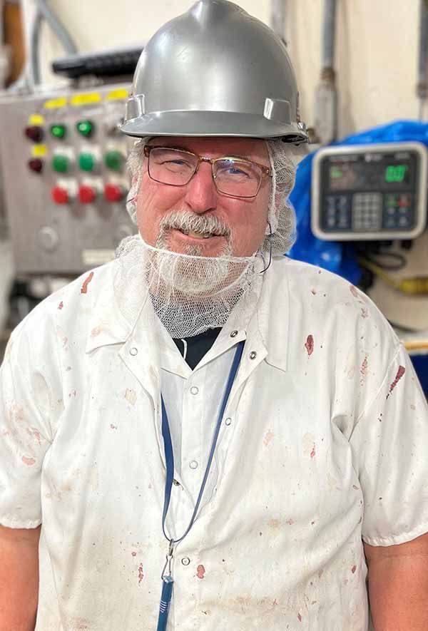 A worker in a gray hard hat, protective hair net, and white, stained coat stands in a factory or food processing plant.