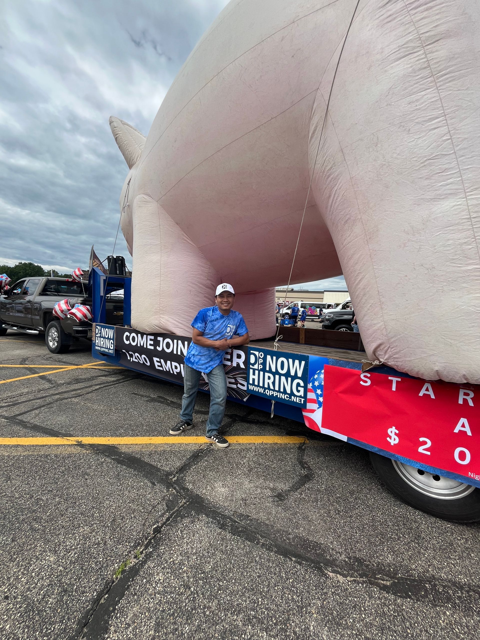 A person stands by a float featuring a large inflatable pig, advertising job openings and a starting pay of $20.