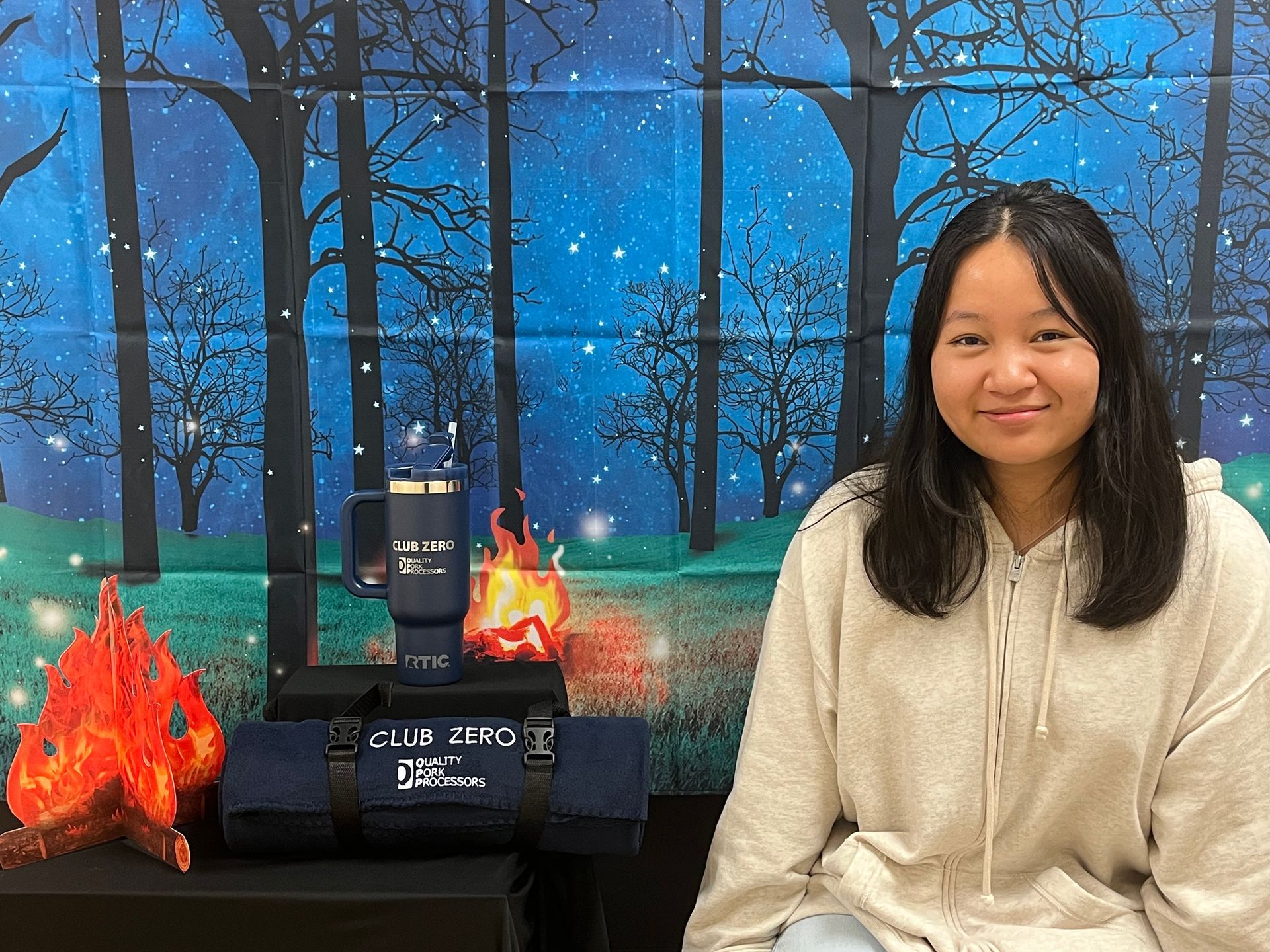A person sits before a campfire-themed backdrop, with a branded tumbler and rolled-up blanket on a table in the foreground.