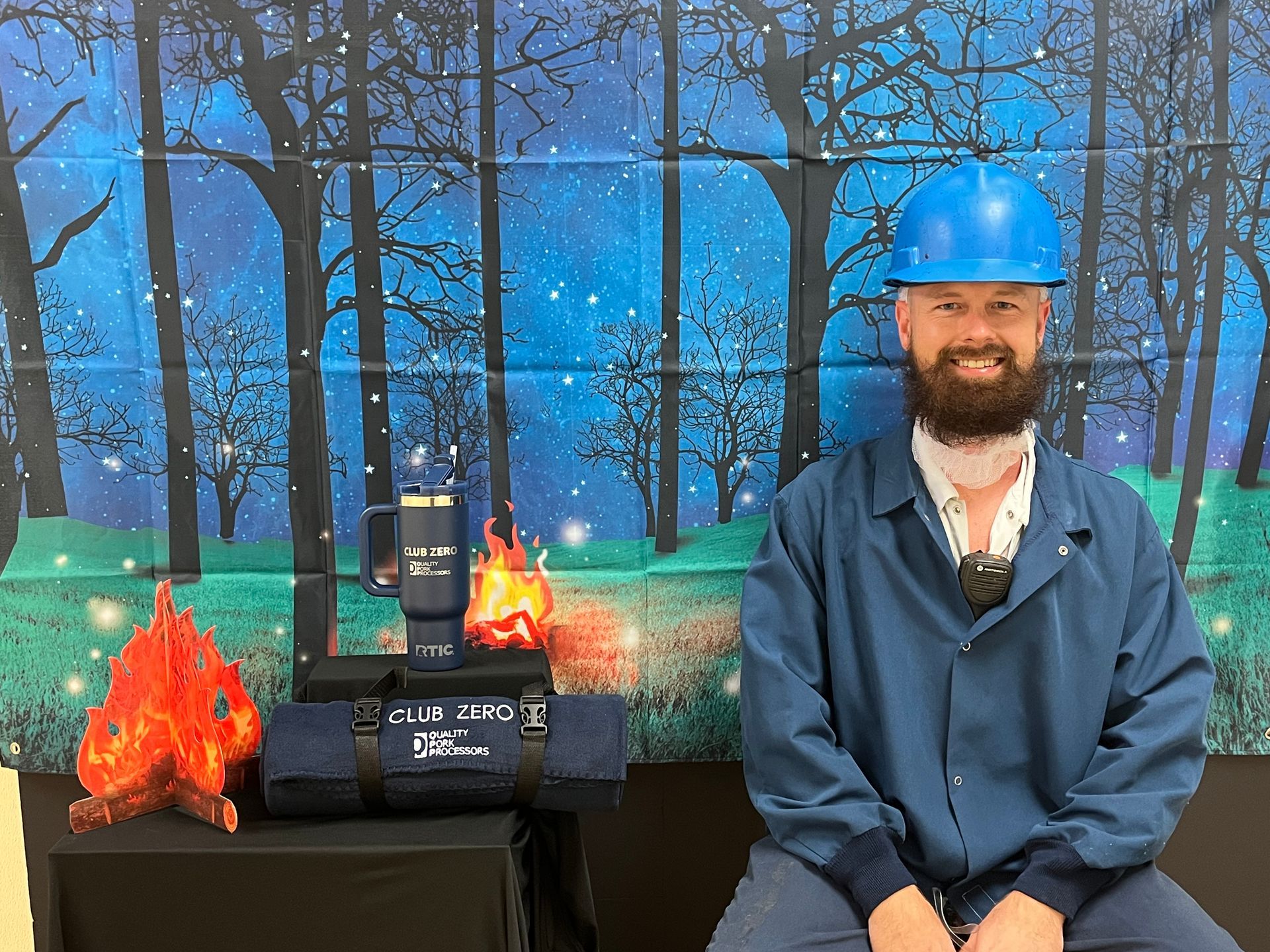 A person in a blue hard hat and work shirt sits next to a campfire prop and a branded camping blanket against a night backdrop.