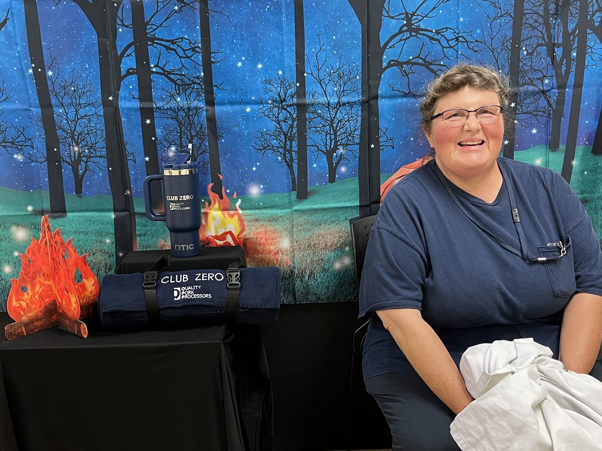A person smiling in front of a forest-themed backdrop, with a tumbler and a folded dark blanket on a black table nearby.