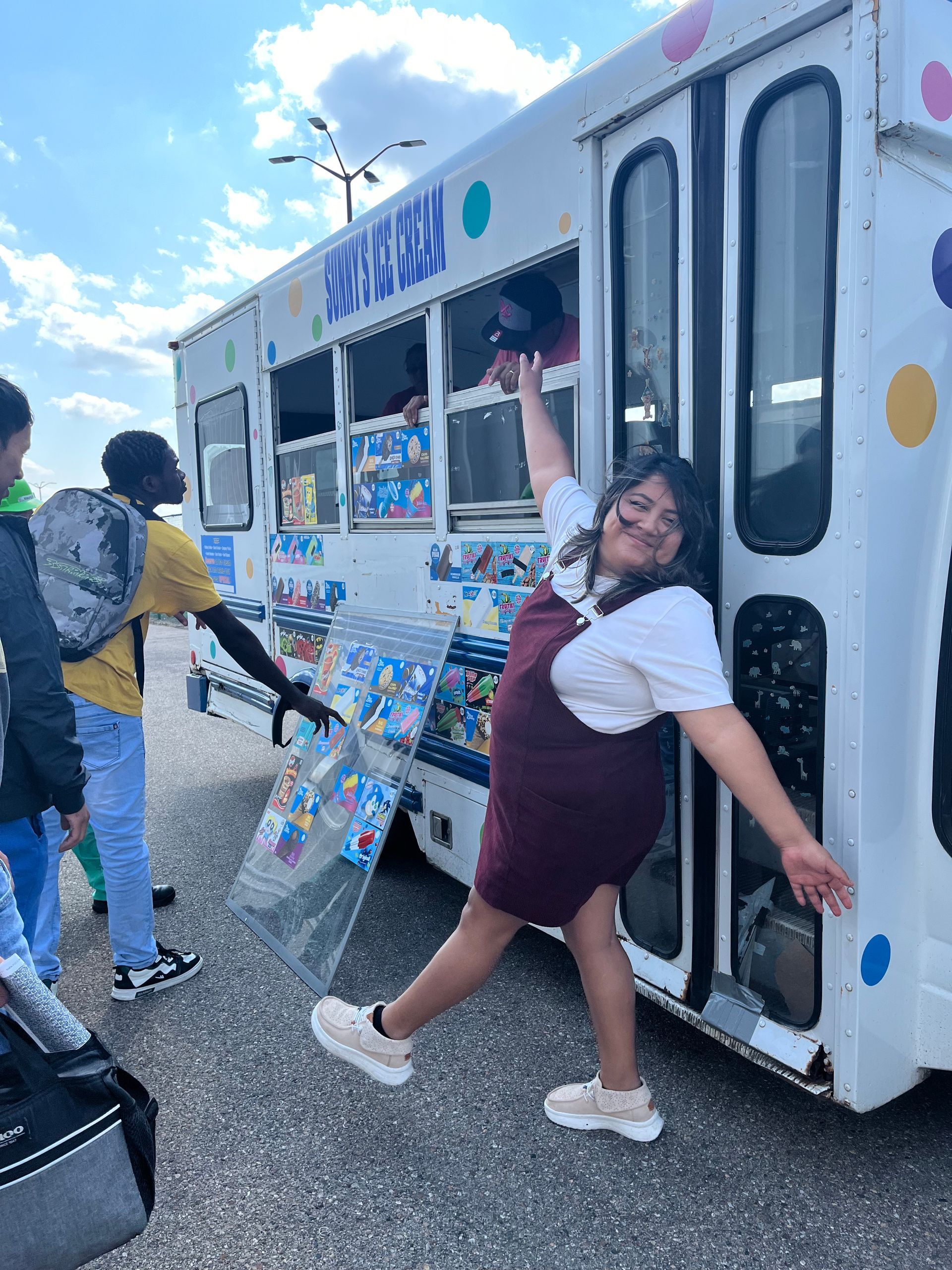 A person wearing a maroon dress and white shirt poses playfully next to an ice cream truck while another person waits.