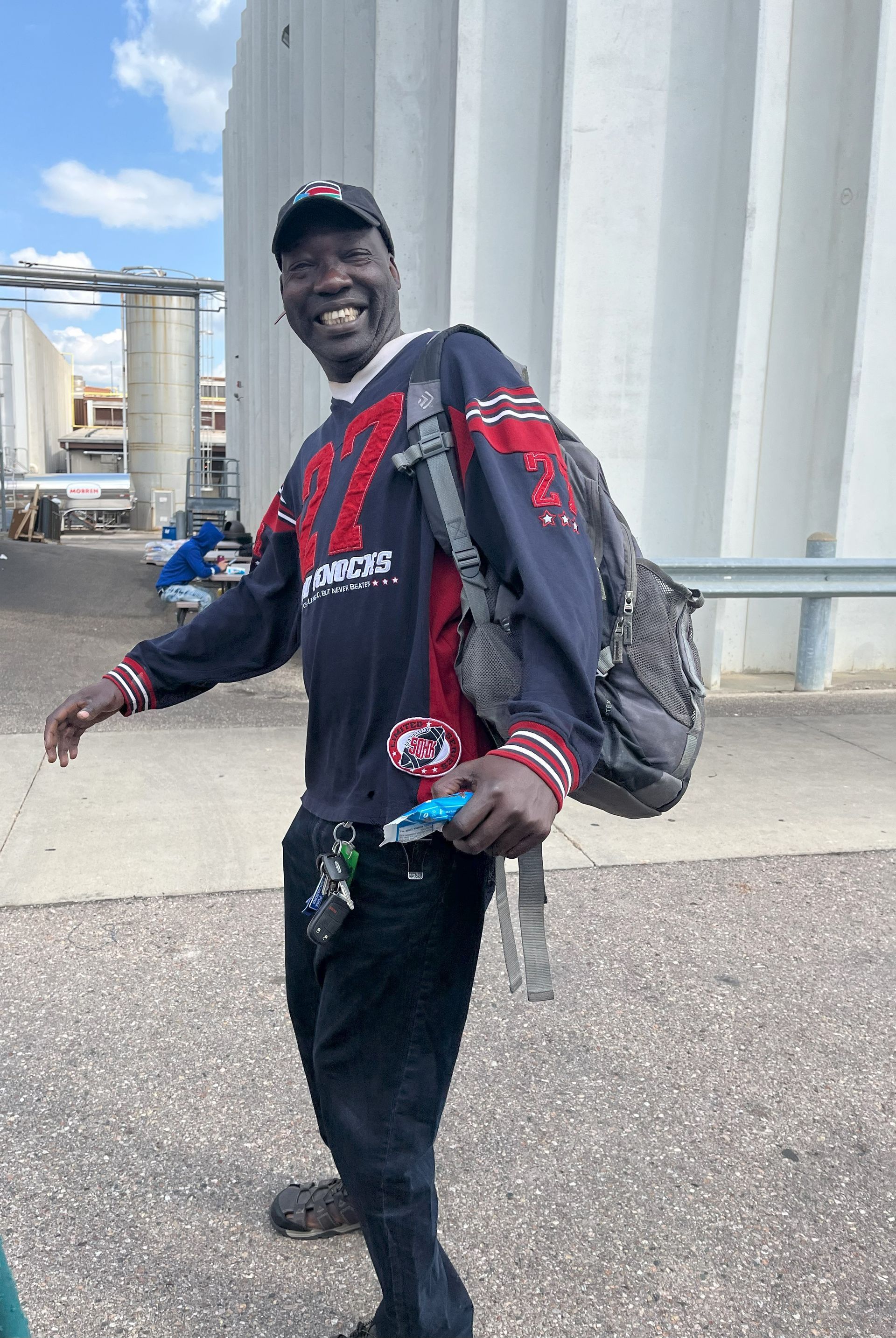 A person wearing a dark sports jersey and a backpack smiles while standing outdoors in front of a large white structure.