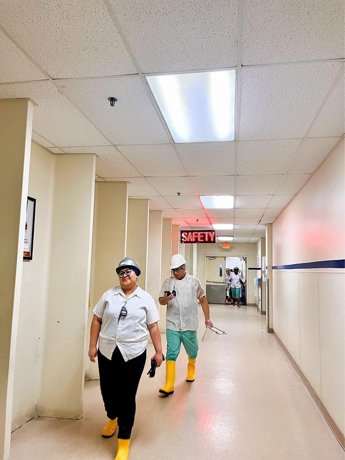 Two people in hard hats and yellow boots walk down a brightly lit office hallway with a red 