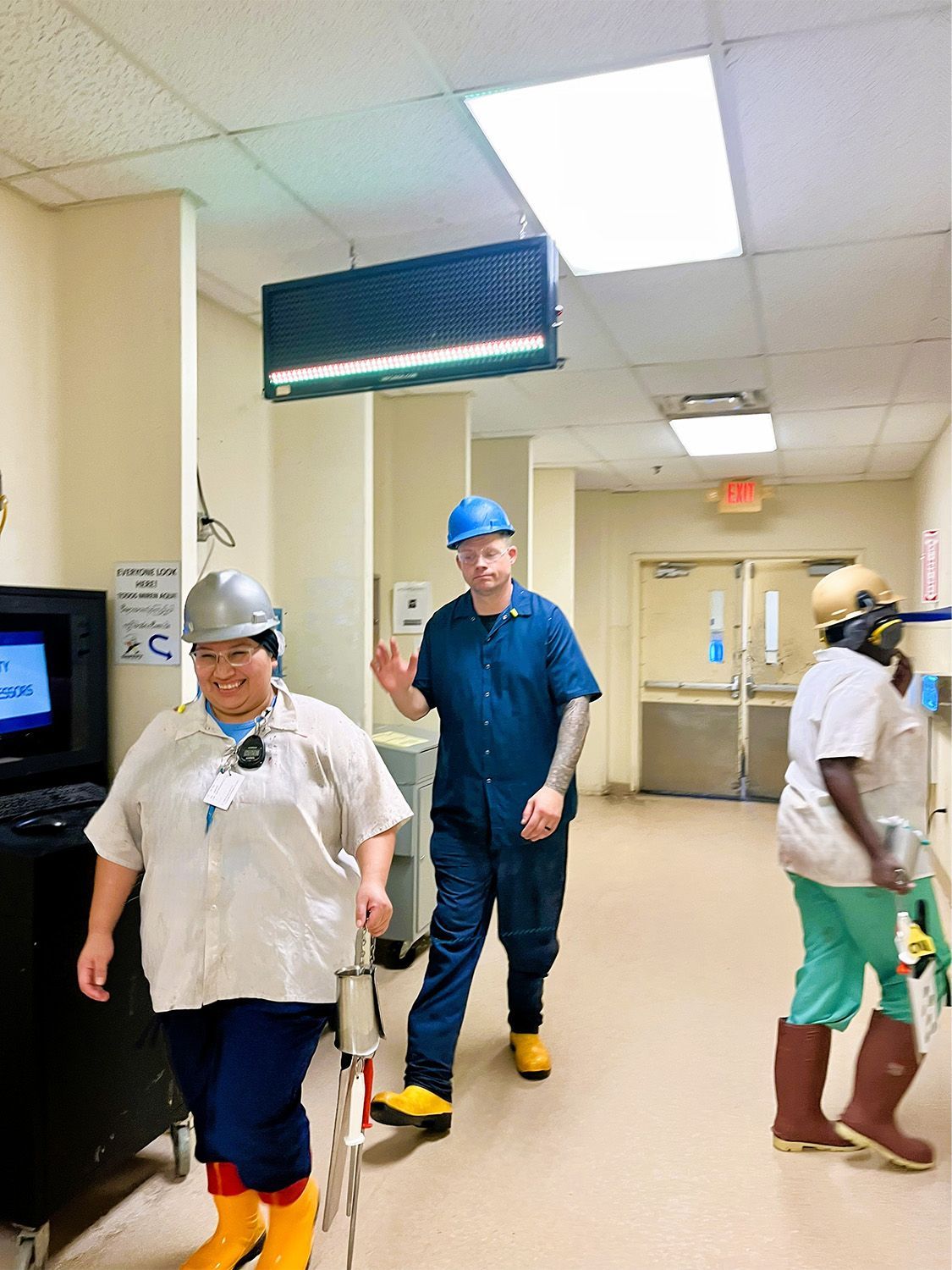 Three people in industrial uniforms and hard hats walk down a brightly lit hallway in a facility.