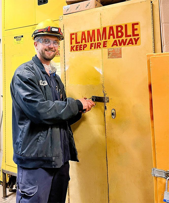 A smiling worker in safety gear stands in a warehouse, locking a yellow cabinet labeled 