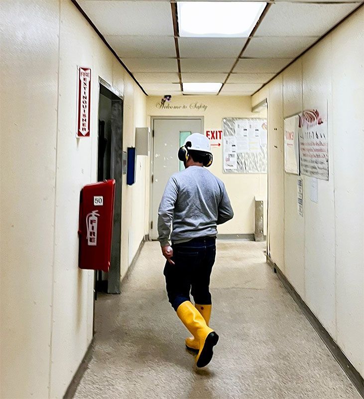 A person in a hard hat and yellow boots walks down a brightly lit industrial hallway past a red fire extinguisher.