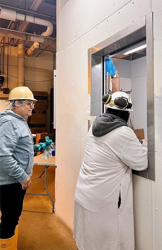 Two people in hard hats and safety gear interact through a service window in an industrial facility.