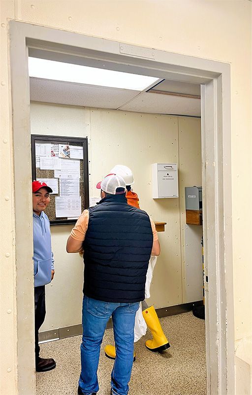 Three people in work attire stand in an office doorway looking at a bulletin board on the wall.
