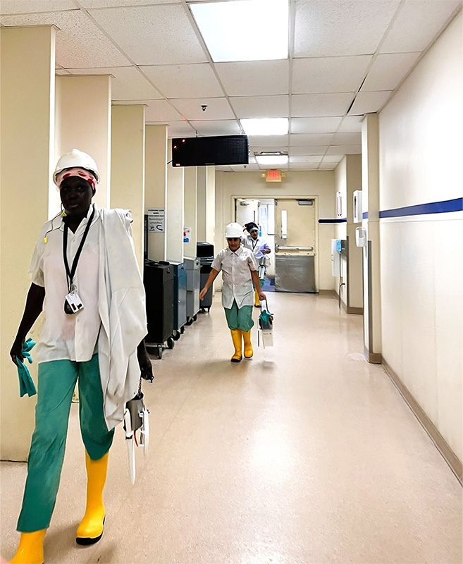 Two cleaners in white hard hats and yellow boots walk down a hospital hallway carrying cleaning equipment.