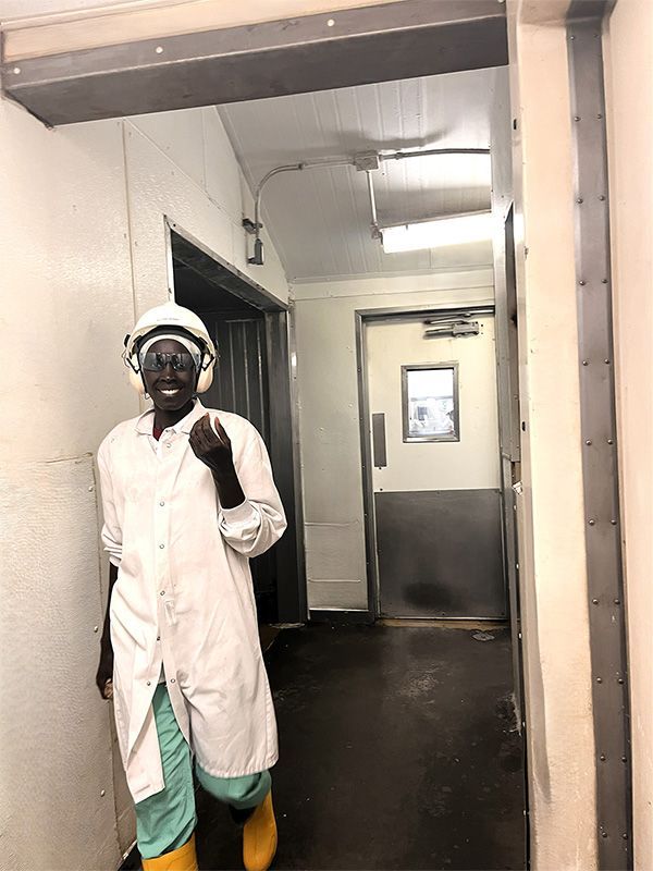 A person in a white lab coat, green pants, yellow boots, and safety gear walks through a hallway toward a metal door.