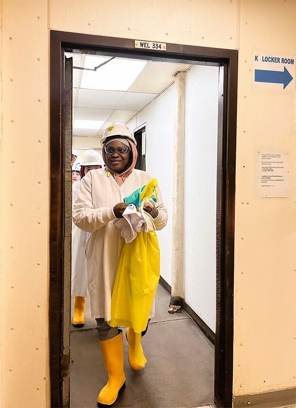 A person in a white lab coat, hard hat, and yellow boots walks through a doorway labeled with a blue locker room sign.