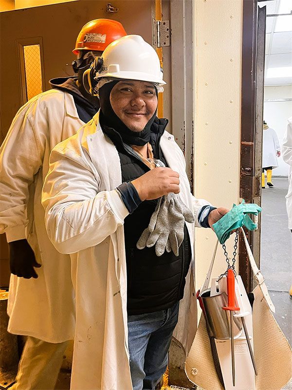 Two workers in white lab coats and hard hats, one smiling while holding protective gloves and equipment.