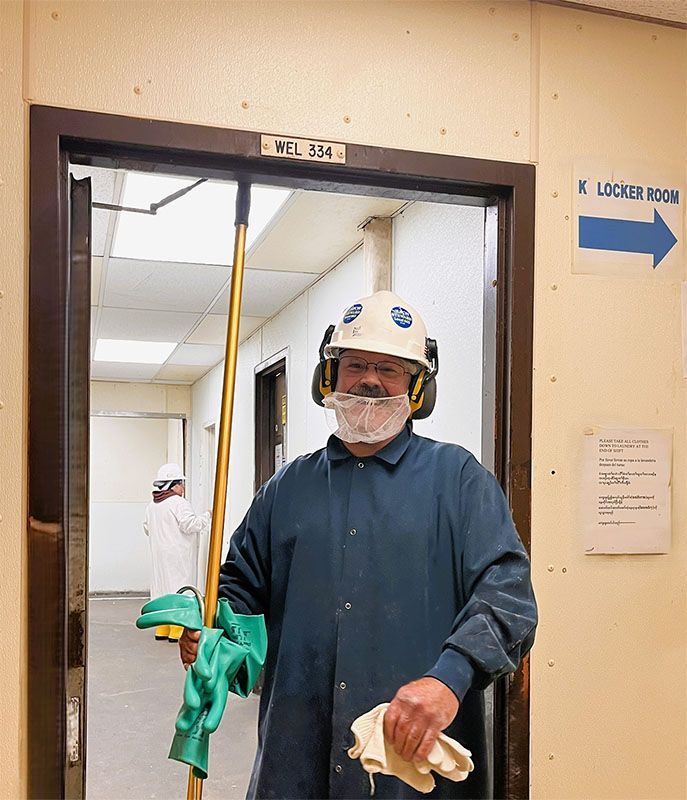 A person wearing protective gear, including a hard hat, ear muffs, and a beard net, stands in a hallway holding gloves.