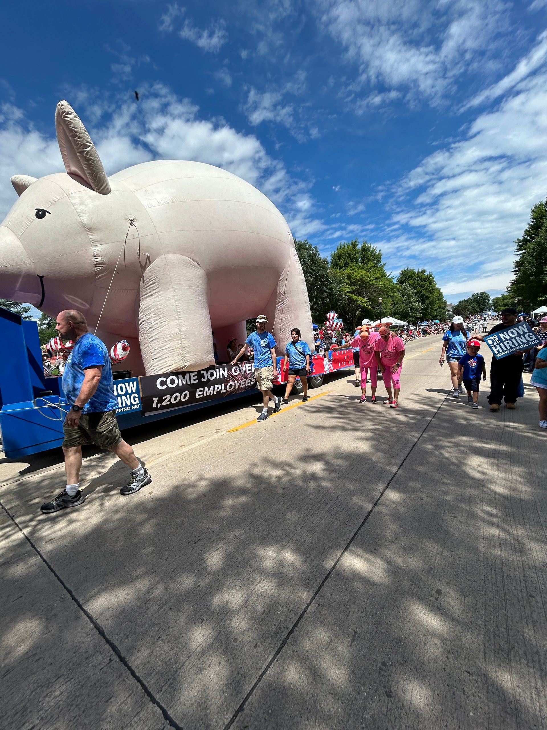 A large inflatable pig on a parade float moves down a sunny street with people walking alongside.