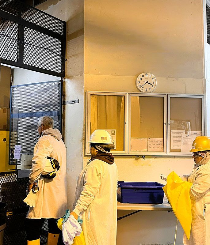 Three people in white protective coats and hard hats stand in a brightly lit industrial workspace.