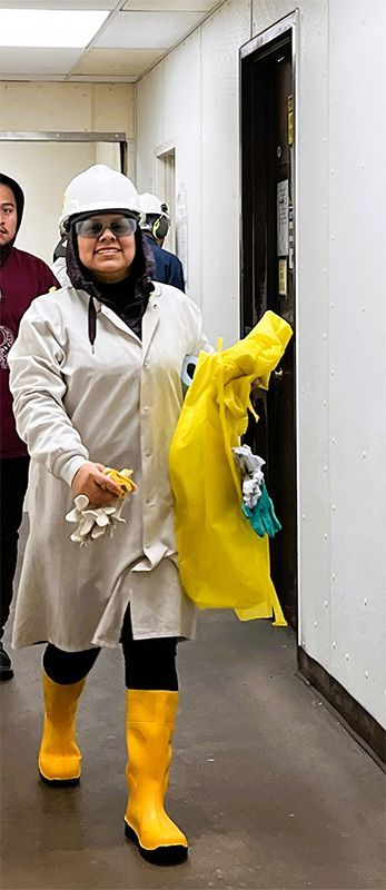 A person in a hard hat, lab coat, and yellow boots walks down a hallway, carrying yellow protective gear and gloves.