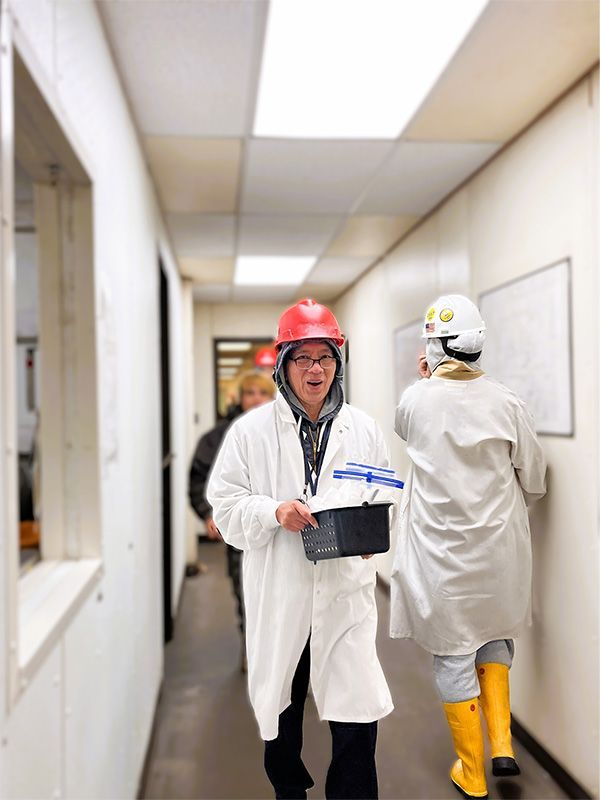 People in lab coats and hard hats walk down a brightly lit industrial hallway.
