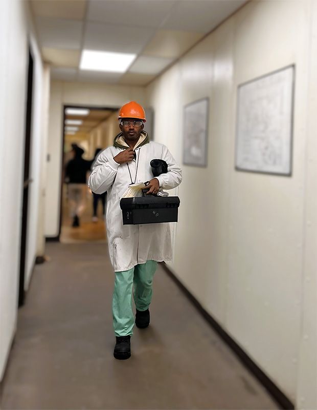 A person in a hard hat, white lab coat, and scrubs walks down a hallway carrying a black box.