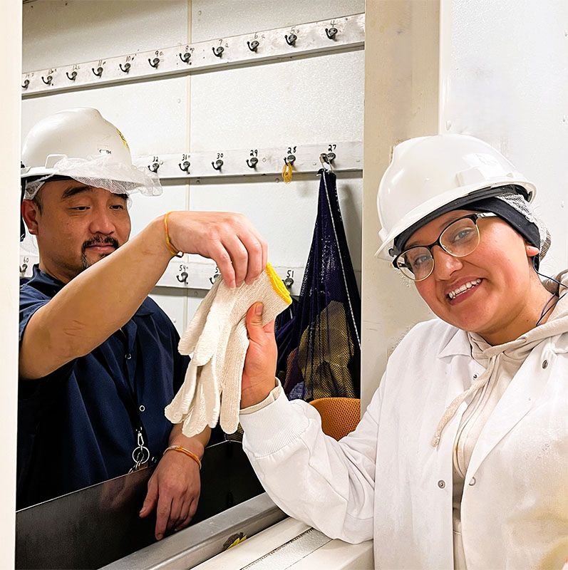 Two people wearing white hard hats and protective gear stand in a workspace, one handing the other a pair of work gloves.