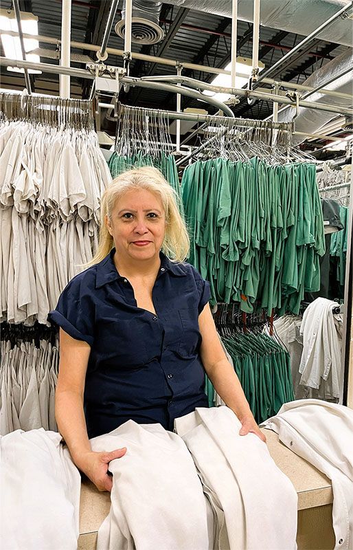 A person wearing a navy shirt stands in a laundry facility, sorting piles of white linens among rows of hanging garments.