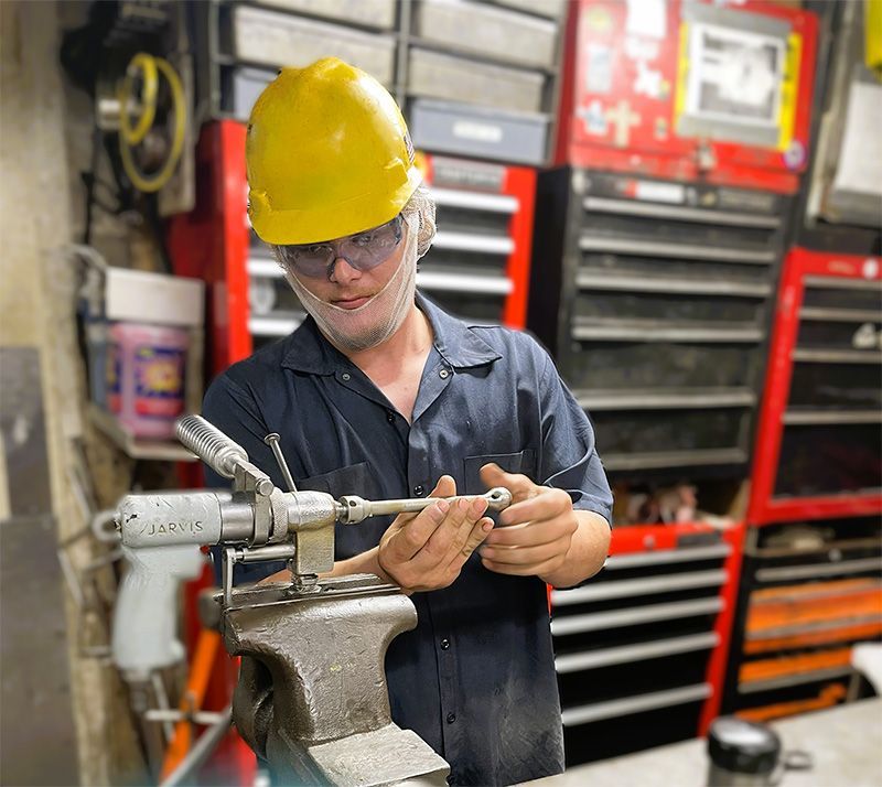 A person wearing a hard hat, safety glasses, and a uniform repairs a metal tool secured in a bench vise in a workshop.
