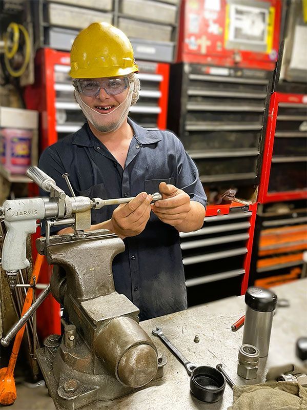 A person in a hard hat and safety glasses works on a mechanical part held in a metal vise at a cluttered workbench.