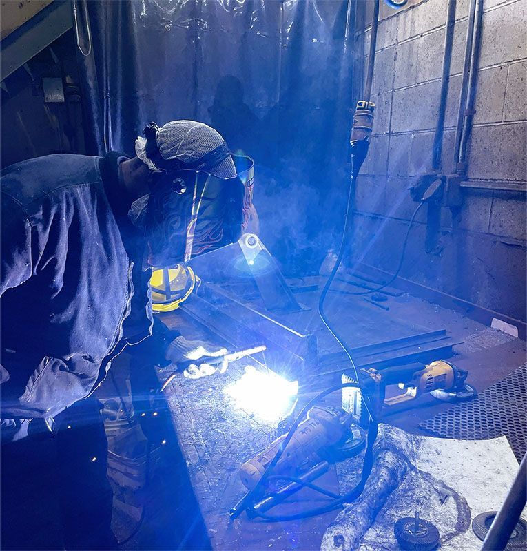 A person wearing a welding helmet performs metal welding, producing a bright blue light and smoke in a workshop setting.