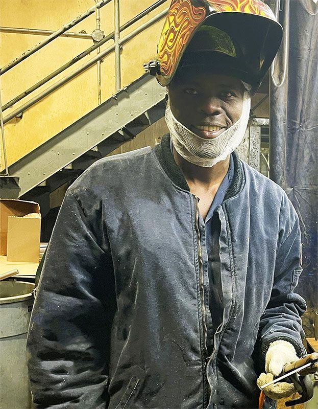 A smiling worker wearing a welding helmet and a protective jacket in an industrial workshop.