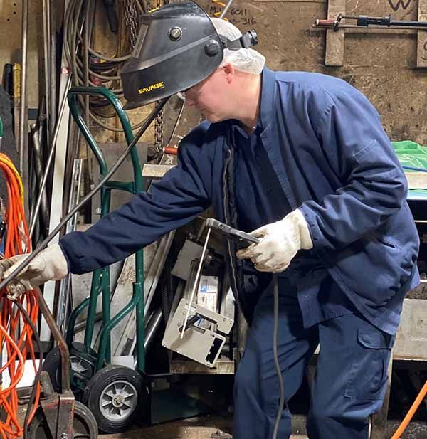 A welder wearing protective gear and gloves holds a welding torch in a cluttered workshop.