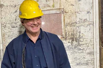 A person wearing a yellow hard hat and dark blue shirt stands smiling in front of a textured, light-colored wall.
