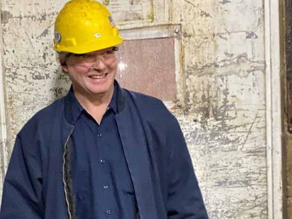 A person in a yellow hard hat and dark blue work jacket smiling in front of a weathered, textured wall.