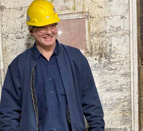 A smiling person wearing a yellow hard hat and a blue jacket, standing in front of a weathered, textured wall.