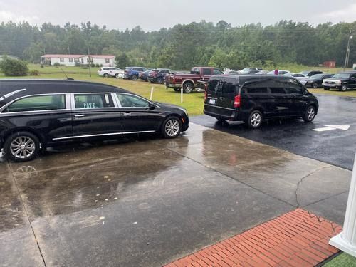 Black hearse and SUV parked on a wet driveway near a grassy field on a rainy day