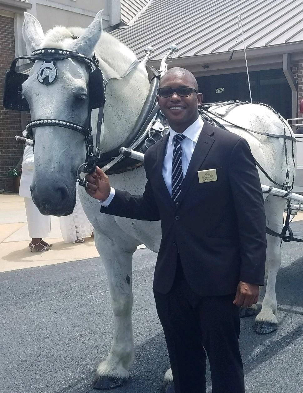 Man in a dark suit standing beside a white horse in front of a building.
