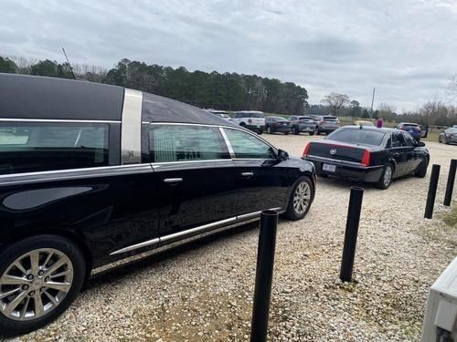 Black car and sedan parked on gravel lot beside black bollards under a cloudy sky.