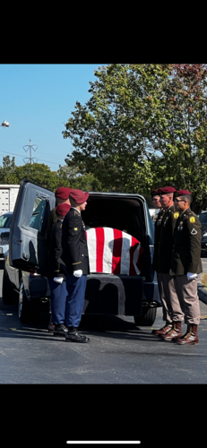 Flag-draped casket in a hearse, with uniformed honor guard standing beside it outdoors.