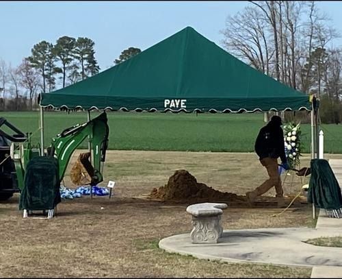 Green canopy tent on a field with workers digging near a sand pile beside a patio and fountain.