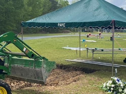 Green tractor loading dirt under a green canopy at a sports field with flowers nearby