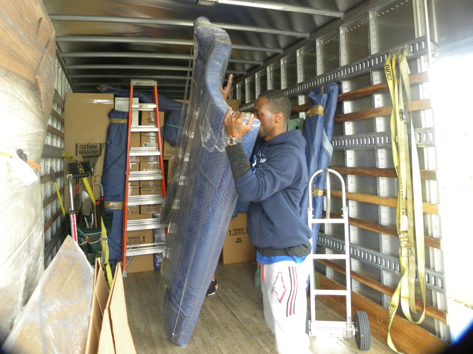 Man standing in a truck trailer, wrapping a large item in protective plastic beside a ladder