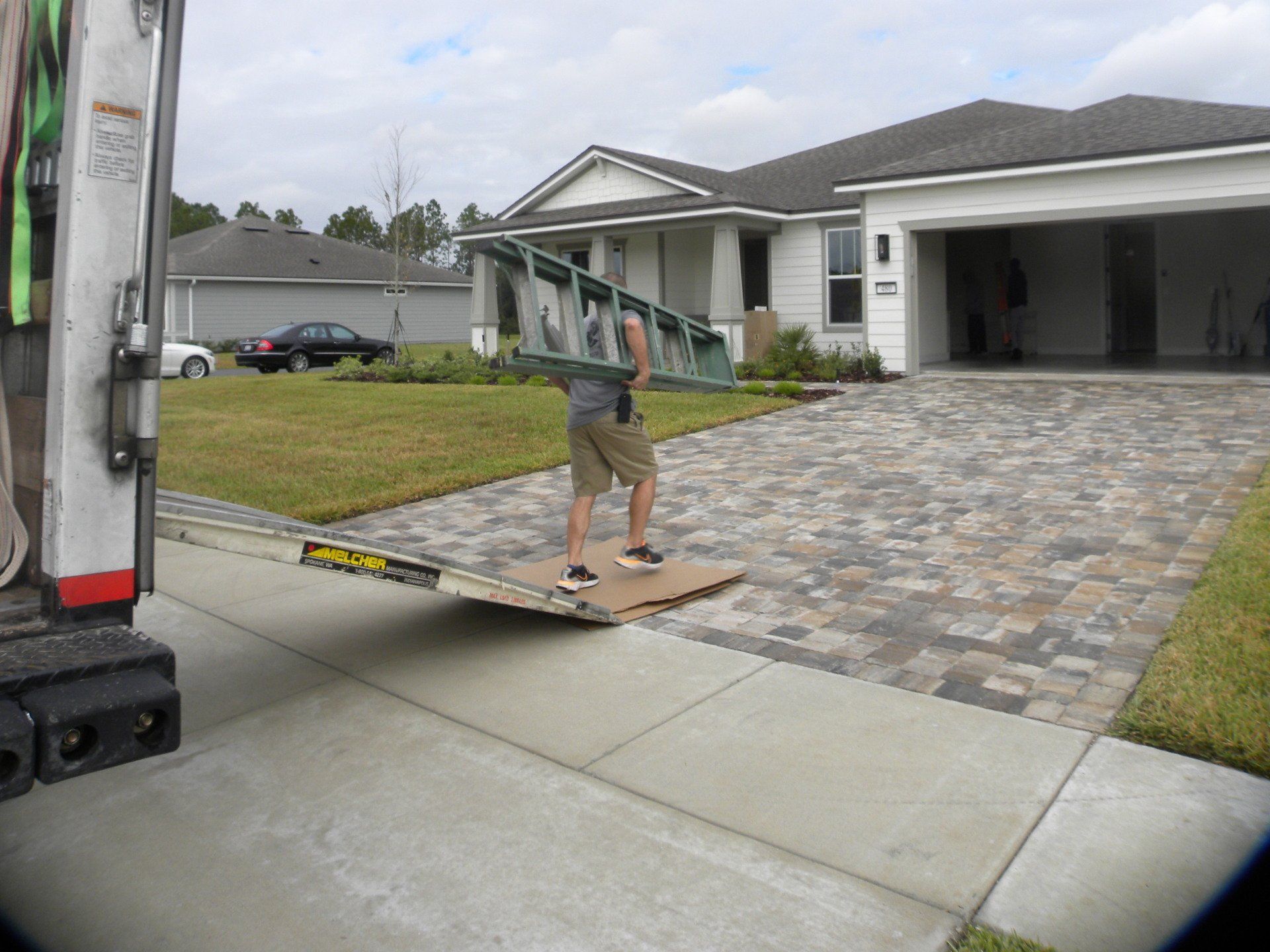 Worker carrying a ladder off a moving truck toward a suburban driveway and garage.