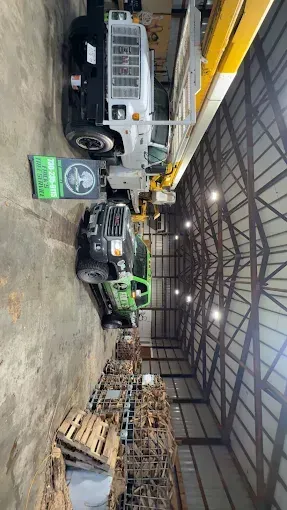 Industrial warehouse with a white truck, forklift, stacked pallets, and a green sign under a metal roof.