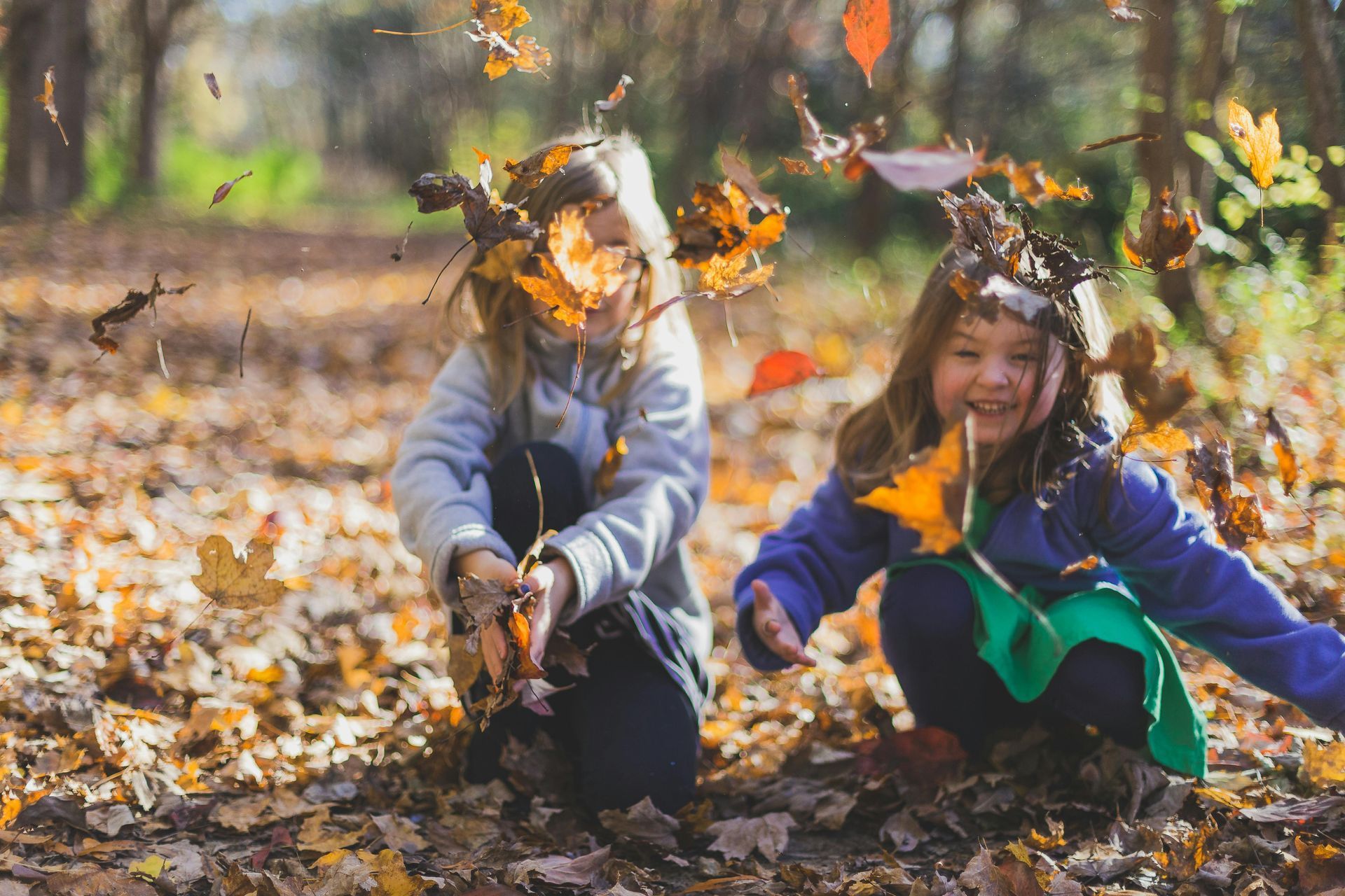 Girl jumping and laughing in a park, throwing autumn leaves; blue sky and trees in background.