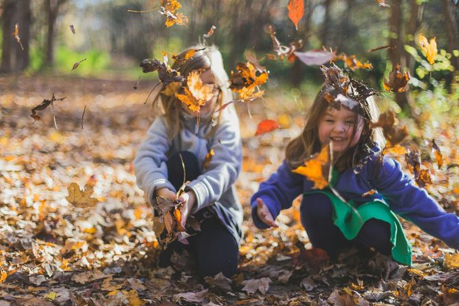Girl jumping and laughing in a park, throwing autumn leaves; blue sky and trees in background.