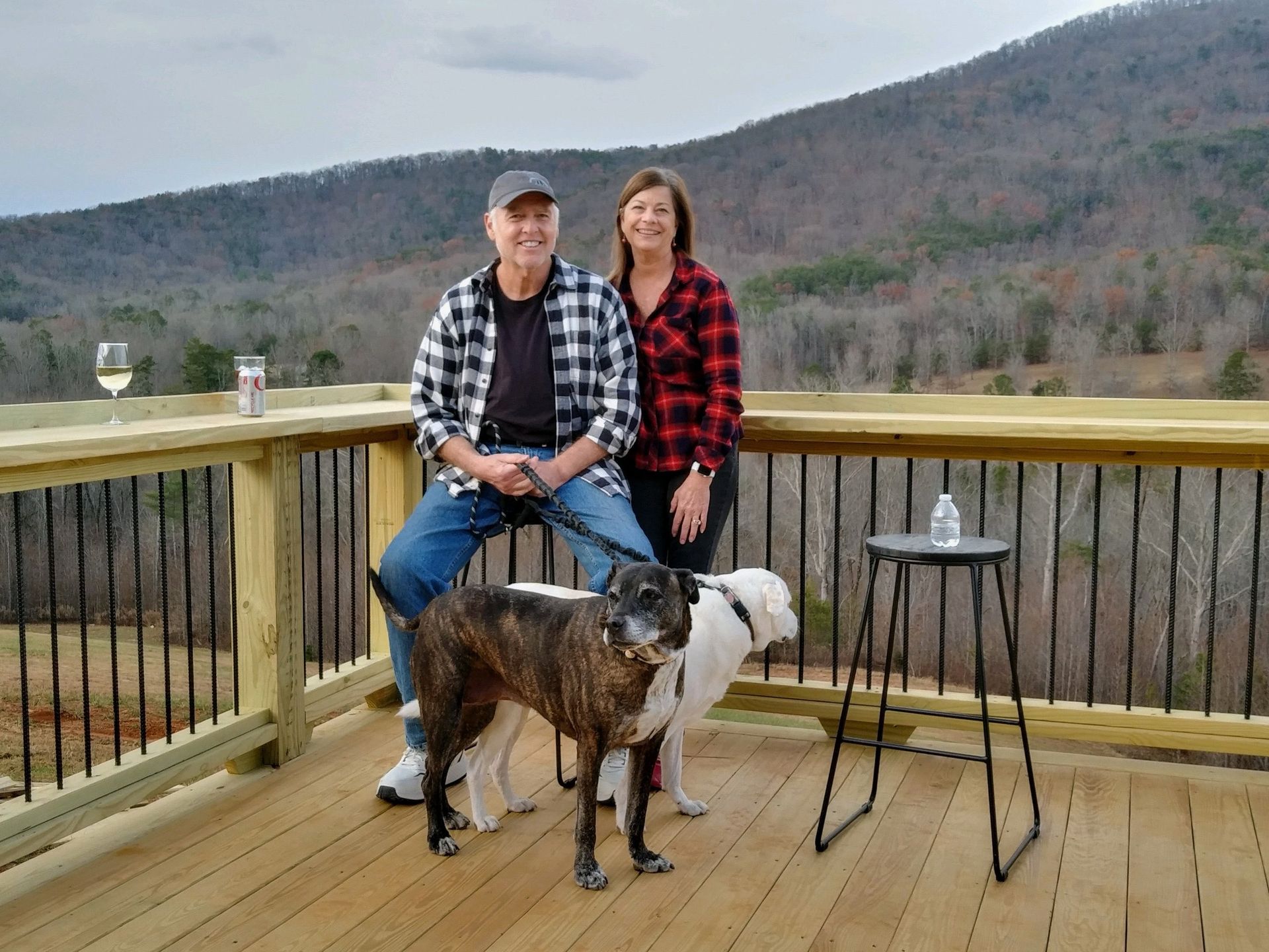 Man and woman on a deck with a brindle dog, mountains in the background, beside a small table