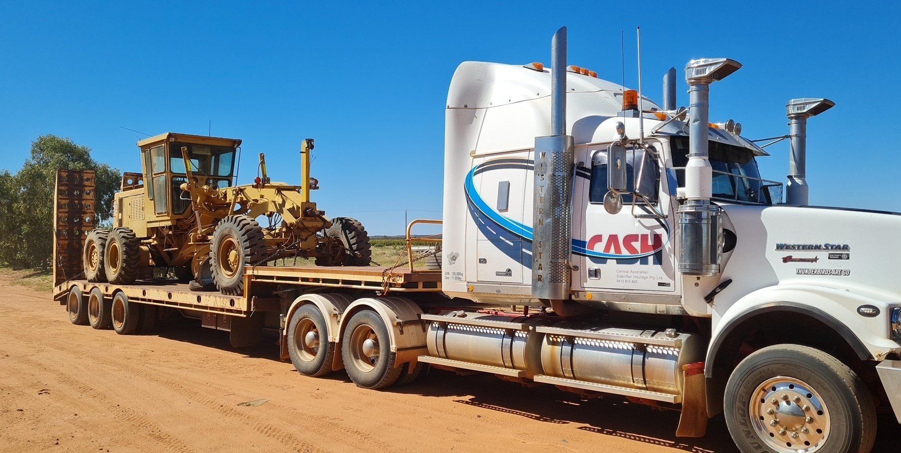 A Yellow Grader on A Flatbed Trailer Being Pulled by A Semi-Truck — Central Australian Sidelifter Haulage in Ciccone, NT